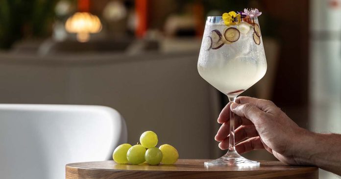 Mão segura um copo de cocktail decorado com flores e rodelas de fruta, numa imagem associada ao EVOLUTION Bar Fest