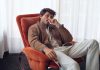 Moda masculina com linhas urbanas e cortes descontraídos na nova coleção da OOTO Moda masculina OOTO com blusão castanho, malha clara e calças em tom cru, num homem sentado numa poltrona laranja