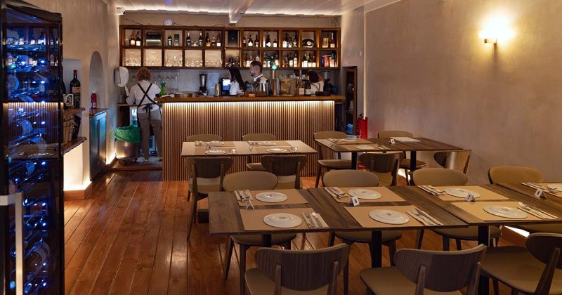 Perspetiva da sala de jantar do restaurante Vizinhos do Rei em Odivelas com garrafeira vertical iluminada em primeiro plano e balc&atilde;o ao fundo.