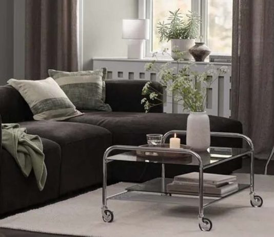 Decoração de outono da JYSK conjuga o brilho dos cromados com o conforto dos tons castanhos Sala com sofá escuro, mesa de centro em vidro e cromado com rodas e suporte de televisão na decoração de outono da JYSK