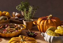 Tachos e panelas Le Creuset levam as formas e as cores do outono à mesa Tachos e panelas Le Creuset em forma de abóbora, em laranja, amarelo e branco, sobre uma mesa com legumes e ervas aromáticas