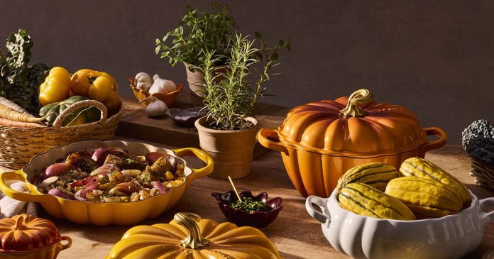 Tachos e panelas Le Creuset em forma de abóbora, em laranja, amarelo e branco, sobre uma mesa com legumes e ervas aromáticas